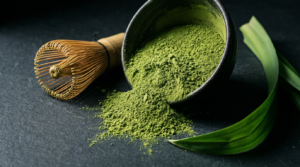 Jade-green pandan matcha powder in a dark bowl with bamboo whisk and fresh pandan leaf