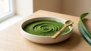 A shallow ceramic bowl of fine vivid green pandan powder with a spoon swirl drawn through it and a bamboo spoon resting on the rim